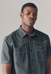 Camicia a maniche corte di colore grigio antracite realizzata in tessuto strutturato. Presenta due tasche sul petto, chiusura con bottoni e cuciture bianche a contrasto.