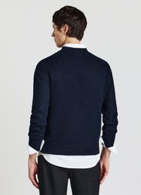 Mann in einem dunklen marineblauen Pullover über einem weißen Hemd mit schwarzen Hosen, steht mit dem Rücken zu einer schlichten hellen Hintergrund.