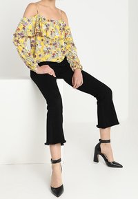 Blusa gialla floreale off-shoulder con dettagli a volant, abbinata a pantaloni neri a vita alta a gamba svasata e scarpe con tacco nere con cinturini alla caviglia.