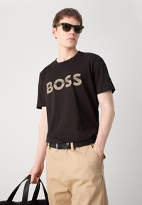 Melna kokvilnas T-kreklu ar "BOSS" logo pelēkā krāsā, kombinēts ar brūniem biksēm, melnu jostu un tumšiem saulesbrillēm. Modelis tur melnu somu.