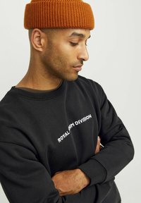 Felpa nera con il testo bianco "ROYAL DENIM DIVISION" sul petto; abbinata a un berretto di maglia arancione testurizzato.