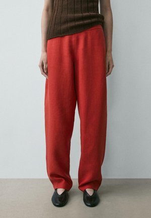 Persona che indossa pantaloni rossi larghi, scarpe nere senza lacci e un top marrone senza maniche a coste, in piedi contro un muro grigio semplice.