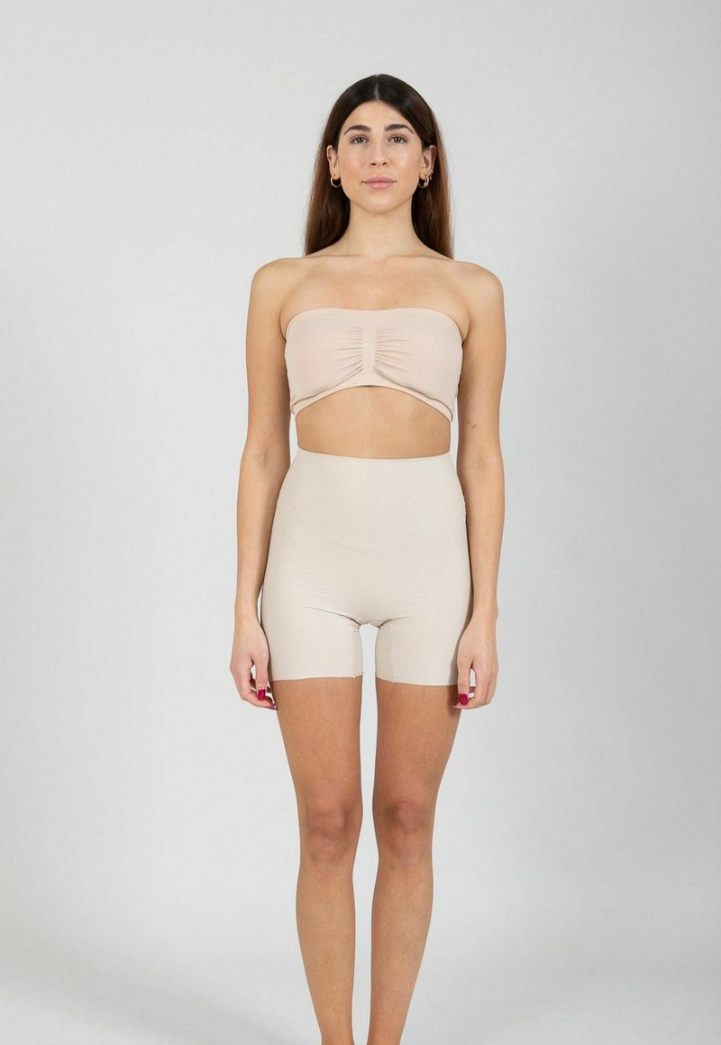 Femme debout de face portant un haut bandeau nude sans bretelles et un short cycliste taille haute nude sur un fond gris uni.