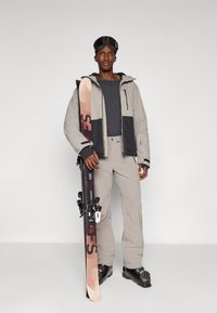 Ski outfit featuring a beige and black insulated jacket, gray top, and light gray pants. Holding black and pink skis with bindings.