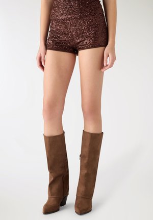 Donna con pantaloncini di paillettes marroni e stivali di camoscio marroni alti al ginocchio, in piedi davanti a uno sfondo bianco semplice.