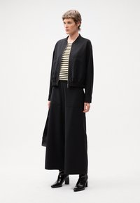 Veste bomber noire zippée sur un t-shirt rayé, assortie avec un pantalon large noir et des bottines à talons noires, présentée dans un cadre minimaliste.