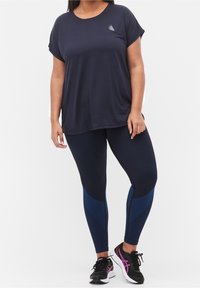 Kobieta ubrana w luźną granatową koszulkę i granatowe legginsy z niebieskimi wzorzystymi panelami, stoi z jedną ręką swobodnie opuszczoną wzdłuż boku.