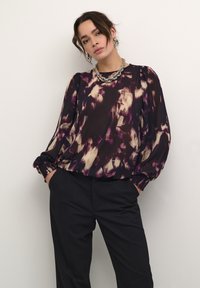 Flödande blus med långa ärmar i lila och beige abstrakt mönster; mjukt material med smockade mudd; kombinerad med svarta, skräddarsydda byxor.