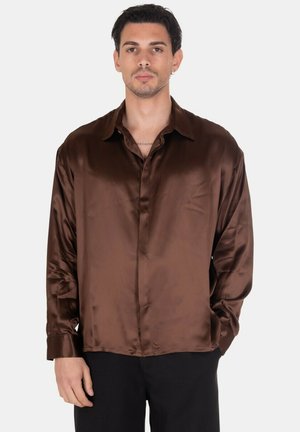 Uomo che indossa camicia marrone lucida a maniche lunghe con colletto e bottoni, pantaloni neri, in piedi davanti a uno sfondo chiaro uniforme.