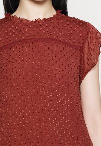 Blusa de color óxido con un patrón de puntos texturizados, cuello con volantes y mangas cortas transparentes. Tela ligera con un acabado suave y sedoso.