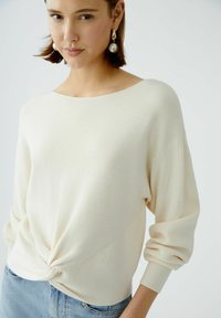 Off-white geripptes Strickpullover mit lockerer Passform, Bootsausschnitt und gerafftem Knotendetail an der Taille; kombiniert mit hellblauen Jeans.