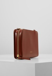 Braune Lederhandtasche mit rechteckiger Form, ausgestattet mit einem Reißverschluss in Goldoptik und einer kleinen Anbringung. Glatte Textur, minimalistisches Branding.