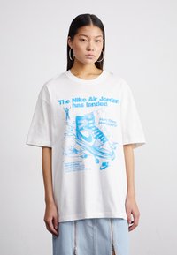 Valkoinen ylisuuri t-paita, joka on valmistettu puuvillasta, sisältää sinisen graafisen suunnittelun lenkkarista sekä tekstiä Nike Air Jordanista, joissa on lyhyet hihat.