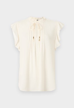 Blouse sans manches couleur crème avec col haut à volants, lien à nouer avec accents dorés, et délicats plis sur le devant.