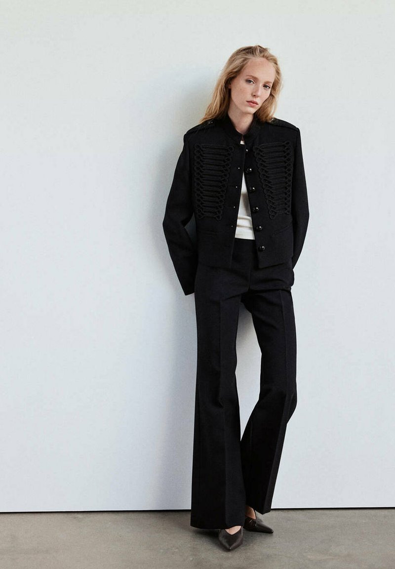 Femme aux longs cheveux blonds portant une veste noire de style militaire, un pantalon noir évasé, un haut blanc et des chaussures noires à bouts pointus, debout contre un mur blanc.