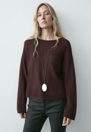 Donna con capelli biondi lunghi che indossa un maglione largo color bordeaux con una tasca sul petto, pantaloni neri e una lunga collana nera con un pendente bianco.