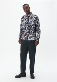 Camisa de manga larga abotonada en blanco con patrones abstractos en azul y marrón; combinada con pantalones oscuros y holgados y zapatos negros.