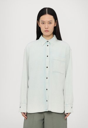 Lys blå denimskjorte med lange ermer, knappestolpe foran og en enkelt brystlomme. Har kontrasterende knapper og klassisk krage.