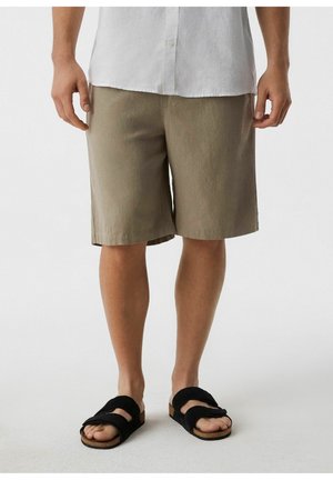 Mężczyzna w beżowych szortach do kolan, białej koszuli i czarnych sandałach z dwoma paskami, stojący na jednolitym tle.