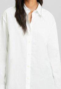 Camicia bianca con bottoni realizzata in un tessuto leggero, caratterizzata da un colletto a punta, maniche lunghe e una linea dritta con una texture sottile.