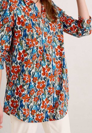 Lang blomstret bluse med røde, blå og grønne blomster, båret af en person med lyse bukser mod en ensfarvet baggrund.