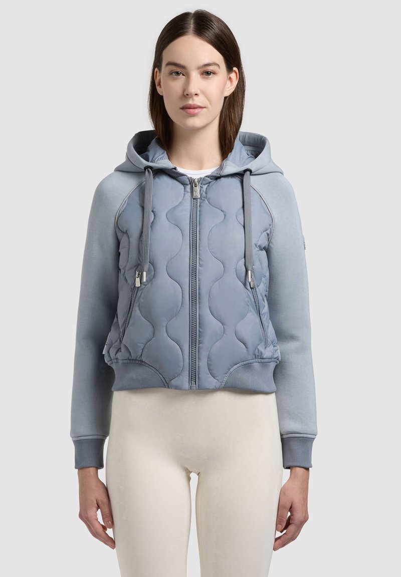 Jeune femme portant un sweat à capuche matelassé bleu clair avec fermeture éclair et cordons, ainsi qu'un pantalon crème, debout devant un fond gris uni.