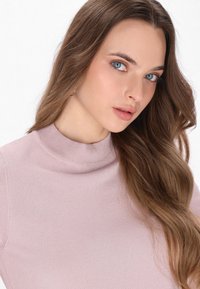 Světle růžový rolák s měkkou texturou a mírným leskem. Modelka má dlouhé vlnité hnědé vlasy a modré oči.