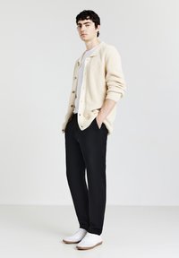 Gilet en maille beige avec de grands boutons, T-shirt blanc, pantalon noir et baskets blanches avec semelles marron, se tenant devant un fond clair.