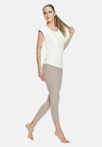 Lys cremet, tætsiddende T-shirt med korte ærmer og rund hals, parret med beigefarvede leggings i ankel-længde. Modellen står barfodet.
