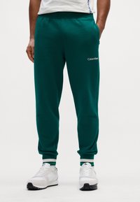 Zöld melegítő nadrág puha anyagból, tapered szabással, amelyen Calvin Klein logó található a combon. Fehér sportcipővel és csíkos mandzsettákkal párosítva.