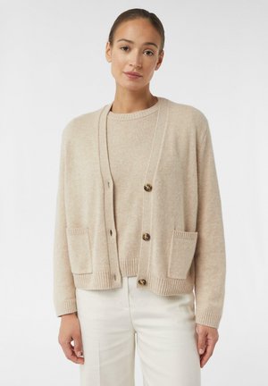 Lichtbeige cardigan gemaakt van zacht materiaal, met vier knopen, geribbelde mouwen en twee voorzakken. Gecombineerd met een bijpassende trui.