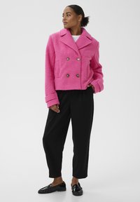 Veste rose floue à double boutonnage avec grand col, poches avant et boutons argentés, assortie à un pantalon noir et des mocassins brillants.