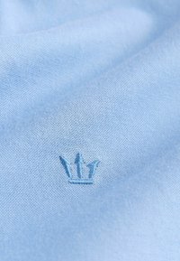 Šviesiai mėlynas audinys su lygiu paviršiumi, kuriame yra mažas sutvirtintas karūnos logotipas atitinkančia spalva.