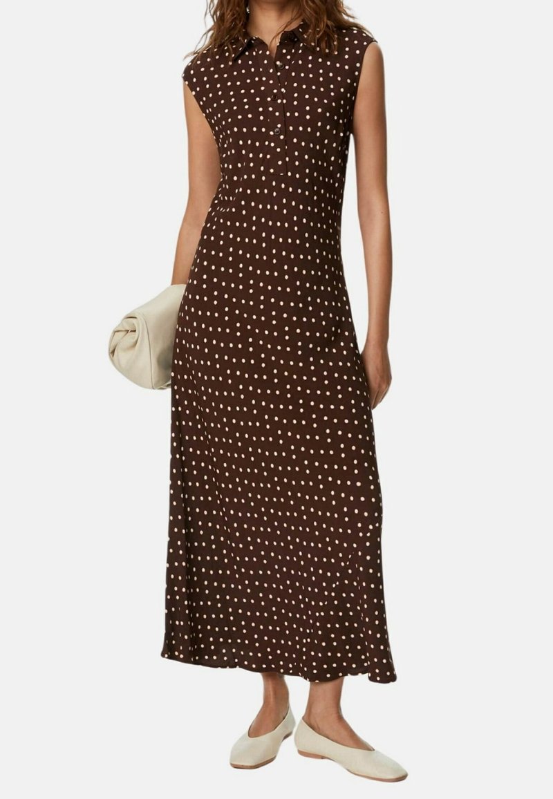 Donna che indossa un abito senza maniche marrone con pois bianchi, tiene in mano una clutch beige e indossa scarpe flat beige su sfondo bianco.