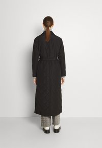 Dlouhý černý prošívaný kabát s páskem v pase, s klopovým límcem, zadním rozparkem a hladkou texturou. Nosený přes kostkované kalhoty a boty.