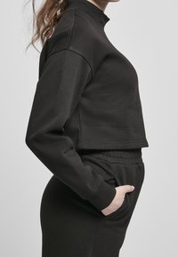 Kvinna i svart långärmad croppad topp och högmidjade byxor med handen i fickan, visad från sidan mot en enfärgad bakgrund.