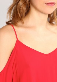 Rød ærmeløs top med V-hals design. Letvægts, flydende stof med en glat tekstur. Tynde stropper og diskret drapering.