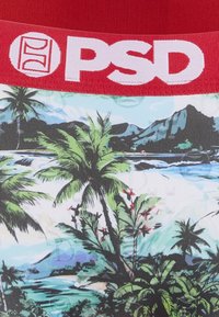 Ceinture rouge avec logo blanc "PSD" au-dessus d'un imprimé tissu représentant des palmiers tropicaux, des fleurs et des montagnes au bord d'un littoral océanique bleu.