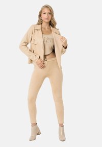 Veste en suede beige ajustée avec un revers et des détails zippés, associée à un haut court assorti et un pantalon skinny. Bottes à talons beiges brillantes.