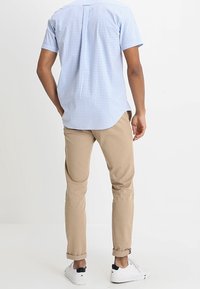 Camicia a maniche corte, in tessuto gingham azzurro chiaro, pantaloni beige, sneakers bianche. La camicia presenta una piega sul retro; i pantaloni sono aderenti con risvolti.