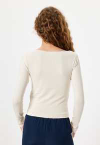 Haut crème à manches longues avec un décolleté droit et un design ajusté. Tissu doux et lisse avec un ourlet légèrement court et sans motifs visibles.