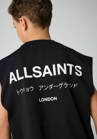 Lühikeste juustega inimene kannab musta ilma varrukateta toppi, millel on valged kirjad "ALLSAINTS", ja jaapani tähed ning "LONDON" seljaosal heleda tausta ees.