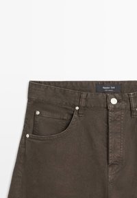 Jean denim marron avec poches avant, rivets argentés, fermeture par bouton, et une étiquette noire portant l'inscription "Massimo Dutti made in Morocco" sur la ceinture.