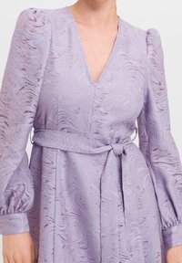 Lavender dress with a V-neck, puffed sleeves, and a cinched waist. Fabric features a textured wave pattern with soft sheen.
