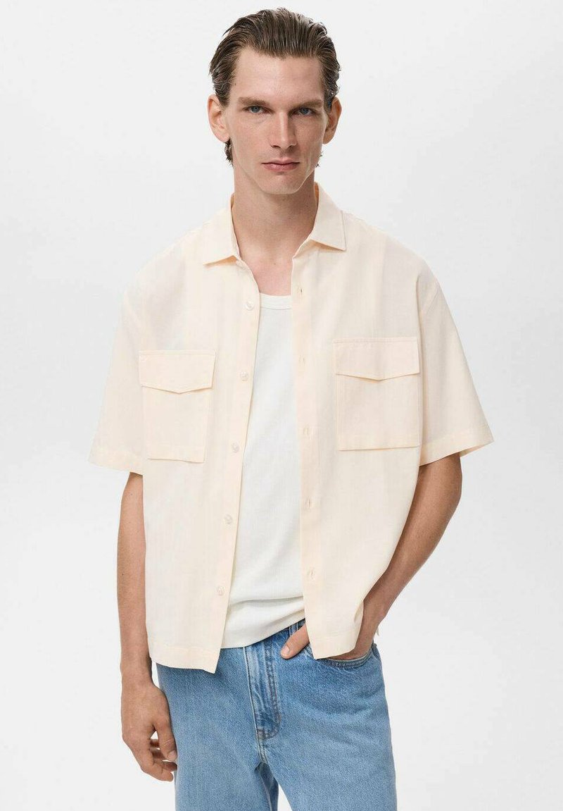 Mand iført en løs kortærmet creamfarvet skjorte med knapper over en hvid tank top og lyseblå jeans, stående med den ene hånd i lommen.