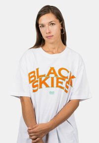 Vit överdimensionerad t-shirt med fet orange text "BLACK SKIES" och gröna accenter. Mjuk bomullstyg, korta ärmar, avslappnad passform.
