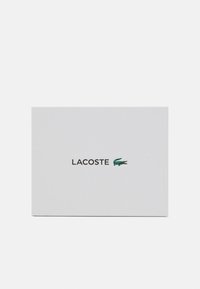 Weißes strukturiertes Kartonpapier mit "LACOSTE" in schwarzem Text und einem grünen Krokodil-Logo rechts. Einfaches und minimalistisches Design.