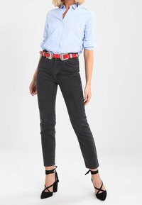 Camisa de cuello azul claro, jeans ajustados gris oscuro, cinturón rojo brillante y zapatos de tacón negros con ataduras al tobillo. Tela suave y ajuste estructurado.