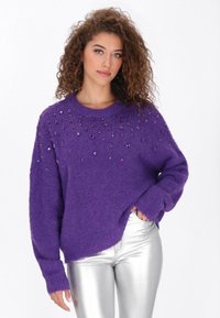 Paarse, fuzzy trui met een ronde halslijn, versierd met paarse edelsteenachtige accenten op de schouders. Gecombineerd met metallic zilveren broek.