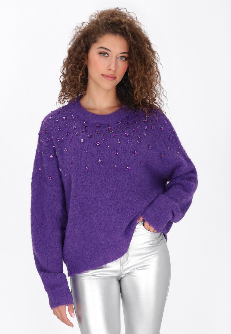 Paarse, fuzzy trui met een ronde halslijn, versierd met paarse edelsteenachtige accenten op de schouders. Gecombineerd met metallic zilveren broek.